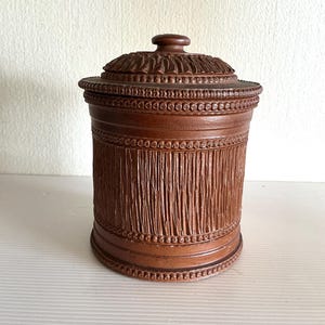 Op de afbeelding: Een bruine, cilindervormige container met een deksel. De container heeft een getextureerd, verticaal patroon en is versierd met decoratieve kralenbanden. Het deksel heeft gesneden details en een kleine knop. Het ontwerp suggereert een vintage of ambachtelijke esthetiek.