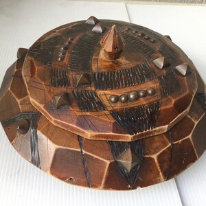 Puede incluir: Una caja de madera con tapa, decorada con un patrón geométrico y tachuelas de metal. La tapa tiene una parte superior puntiaguda y está tallada con detalles intrincados.
