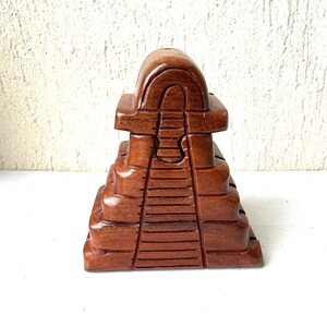 May include: A handcrafted, wooden pyramid-shaped box with a rich brown finish. The box features a stepped design with carved details, resembling ancient architecture. The top has a curved roof-like structure. The box is likely used for decorative or storage purposes.