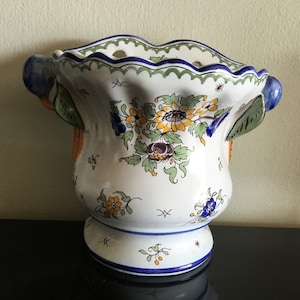 Peut inclure: Un pot en céramique blanche avec des motifs floraux bleus, verts et jaunes. Le pot a un bord festonné et deux poignées avec des accents bleus et verts.