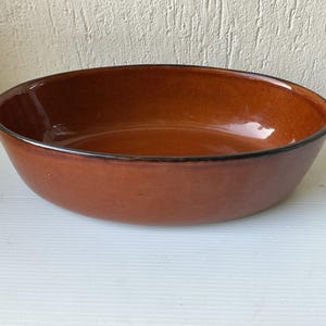 Pode incluir: Uma grande tigela de cerâmica oval. A tigela tem um exterior marrom avermelhado brilhante com uma borda marrom escura. O interior é um tom mais claro de marrom. A tigela está vazia e repousa sobre uma superfície branca.