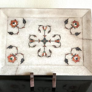 Op de afbeelding: Rechthoekige witte marmeren dienblad met ingelegde bloemmotieven in oranje, groen en zwart. De lade heeft een opstaande rand en wordt ondersteund door twee kleine houten standaards. Het bloemmotief is symmetrisch.