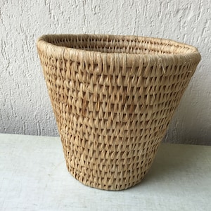 Puede incluir: Una cesta tejida natural hecha de paja con una abertura ancha y redonda y un fondo cónico. La cesta es de color marrón claro y tiene un aspecto natural y rústico.