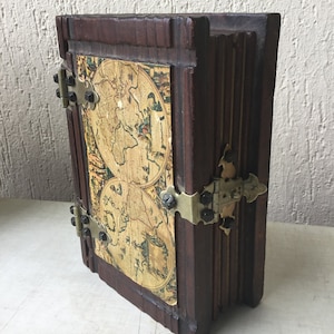 Könnte beinhalten: Eine Holzkiste in Buchform mit einem Vintage-Weltkarten-Design auf dem Deckel. Die Kiste hat Messing-Scharniere und einen Verschluss.