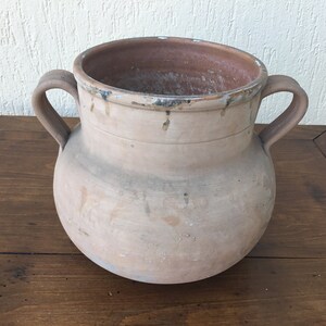 Puede incluir: Una maceta grande de terracota sin esmaltar con dos asas. La maceta tiene un cuerpo redondeado y una abertura ancha. Está hecha de arcilla marrón claro y tiene un aspecto rústico y envejecido.