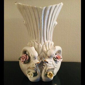 May include: White porcelain vase with two swan figures, each with a pink and yellow flower. The vase has a textured, ribbed design and gold accents.