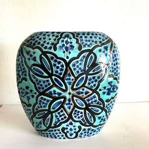 Puede incluir: Un jarrón cuadrado de cerámica con un patrón floral turquesa y azul. El jarrón presenta un diseño simétrico con contornos negros y detalles dorados. La forma general es redondeada con una amplia abertura, adecuada para uso decorativo.