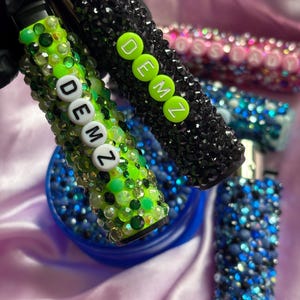 May include: Close-up of several bedazzled lighters in various colours. One is lime green with the word "DEMZ" in white letters. Another is black with the word "DEMZ" in neon green letters. Other lighters are pink and blue.