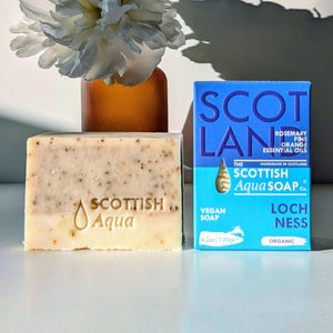 Könnte beinhalten: Eine hellbeige Seife mit dem Schriftzug "SCOTTISH Aqua" in goldener Schrift. Die Seife liegt neben einer blau-weißen Schachtel mit dem Schriftzug "SCOTLAND THE SCOTTISH Aqua SOAP Co. VEGAN SOAP 4.2oz/120g* LOCH NESS ORGANIC" in weißer und goldener Schrift.