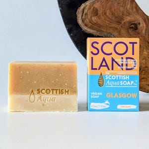 Può includere: Una saponetta Scottish Aqua, beige chiaro alla base e beige più scuro nella parte superiore, accanto a una scatola. La scatola riporta le parole "SCOTLAND" e "SCOTTISH Aqua SOAP Co.", e indica che il sapone pesa 120g. La scatola dice anche "GLASGOW".
