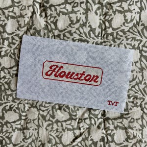 May include: A rectangular white needlepoint canvas featuring the word Houston stitched in red cursive script within a red-bordered rectangular frame. The letters T+T are stitched in red in the bottom right corner of the white mesh fabric.