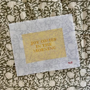 May include: A white needlepoint canvas featuring a rectangular yellow block in the center with the text JOY COMES IN THE MORNING printed in a bold font. The letters T+T are printed in small red ink in the bottom right corner of the canvas.