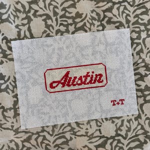 May include: A white needlepoint canvas with the name "Austin" in red script within a red rectangular border. The initials "T+T" are stitched in red in the lower right corner. The canvas is set against a patterned background.