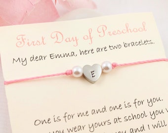 May include: A pink card with the text "First Day of Preschool" and a message to Emma. A pink string bracelet with a silver heart charm with the letter "E" is attached to the card.