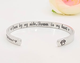 May include: Silver bracelet with the inscription "Once by my side. Forever in my heart" and a paw print charm. The bracelet is on a white background.