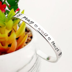May include: Silver bangle bracelet with the words "pray wait trust" engraved on it. The bracelet is decorated with small leaf designs.
