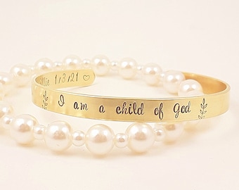 Puede incluir: Un brazalete dorado con la inscripción "I am a child of God" y un diseño floral. Una pulsera de perlas está en el fondo. El brazalete también tiene la inscripción "Ellie 1/3/21" con un símbolo de corazón.