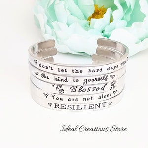 May include: Silver bracelet with four engraved bands. The bands have the following text: "Don't let the hard days win", "Be kind to yourself", "Blessed", "You are not alone", and "RESILIENT".