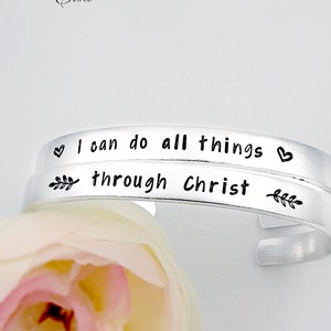 May include: Two silver bracelets with the words "I can do all things through Christ" engraved on them. The bracelets are on a pink flower background.