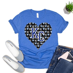 Puede incluir: Una camiseta azul real con un gr&aacute;fico en forma de coraz&oacute;n negro. El coraz&oacute;n est&aacute; lleno de un patr&oacute;n de corazones blancos y las letras "KC" en azul.