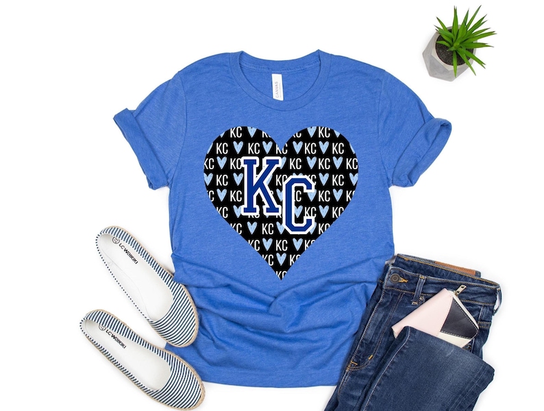 Puede incluir: Una camiseta azul real con un gr&aacute;fico en forma de coraz&oacute;n negro. El coraz&oacute;n est&aacute; lleno de un patr&oacute;n de corazones blancos y las letras "KC" en azul.