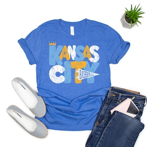 Op de afbeelding: Blauw T-shirt met de tekst "Kansas City Baseball" in een distressed design. De afbeelding bevat een blauwe jeans, gestreepte ballerina's en een kleine plant. Een kleine roze en witte portemonnee is ook zichtbaar.