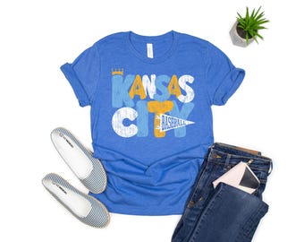 May include: Blue t-shirt with the text "Kansas City Baseball" in a distressed design. The shirt is paired with blue jeans, striped flats, and a small plant. A small pink and white wallet is also visible.