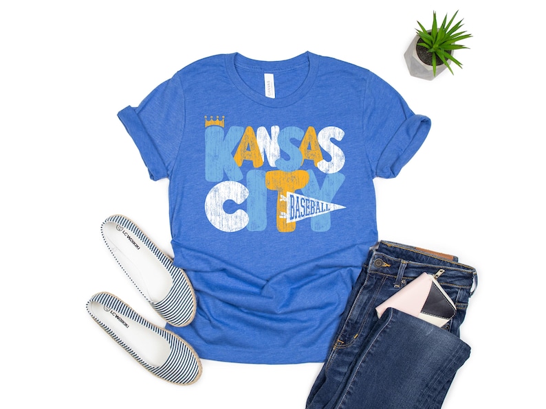 Op de afbeelding: Blauw T-shirt met de tekst "Kansas City Baseball" in een distressed design. De afbeelding bevat een blauwe jeans, gestreepte ballerina's en een kleine plant. Een kleine roze en witte portemonnee is ook zichtbaar.