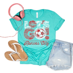 May include: A turquoise blue t-shirt with a distressed white graphic that says "Let's Go! Kansas City" with a red soccer ball in the middle.