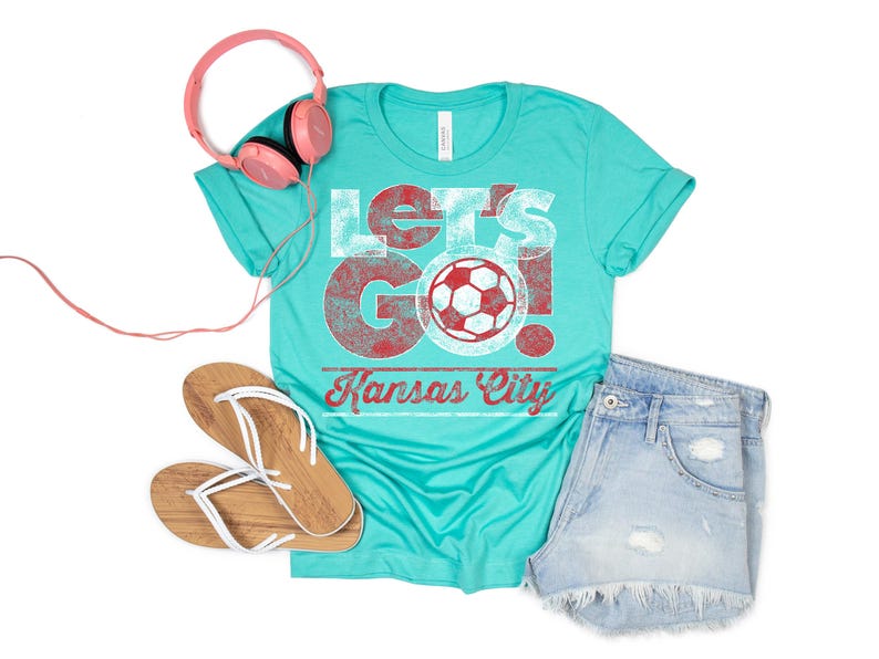May include: A turquoise blue t-shirt with a distressed white graphic that says "Let's Go! Kansas City" with a red soccer ball in the middle.