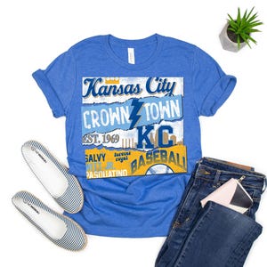 May include: A royal blue t-shirt with a vintage-style graphic design featuring the text "Kansas City Crown Town KC Baseball" with a lightning bolt and a baseball graphic.