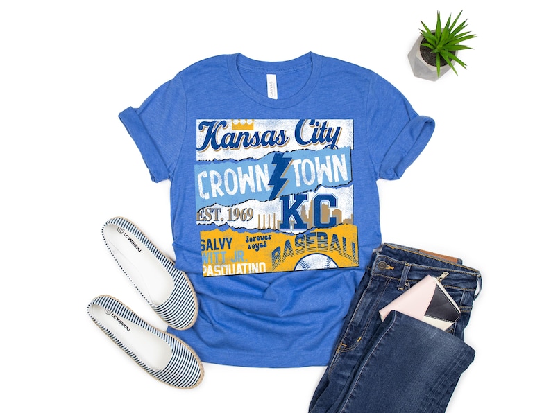 May include: A royal blue t-shirt with a vintage-style graphic design featuring the text "Kansas City Crown Town KC Baseball" with a lightning bolt and a baseball graphic.