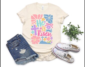 Könnte beinhalten: Ein cremefarbenes T-Shirt mit einem bunten Grafikdesign, auf dem "He is risen" mit Blumen und einem Kreuz steht. Das Shirt hat hochgekrempelte Ärmel und einen Rundhalsausschnitt.