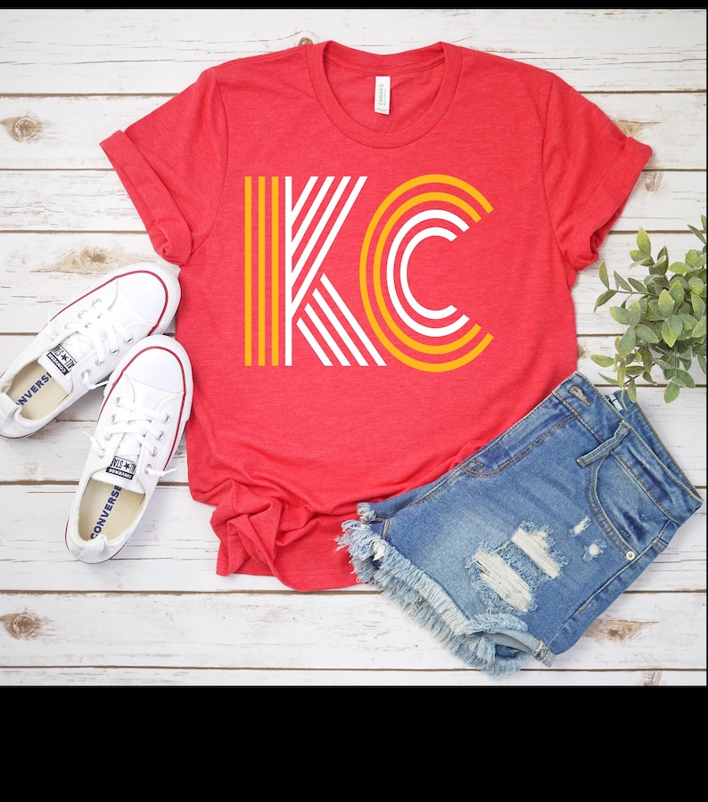 Puede incluir: Una camiseta roja con las letras "KC" en un dise&ntilde;o amarillo, blanco y rojo. La camiseta se combina con pantalones cortos de mezclilla y zapatillas blancas. El conjunto se muestra sobre una superficie de madera blanca.