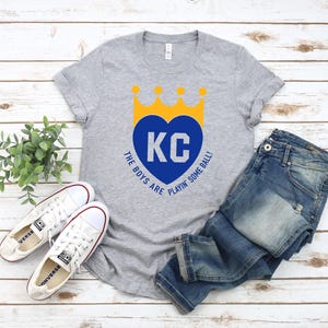 May include: A grey t-shirt with a blue heart and the letters "KC" inside. The heart has a yellow crown on top. The text "The boys are playin' some ball!" is printed below the heart.
