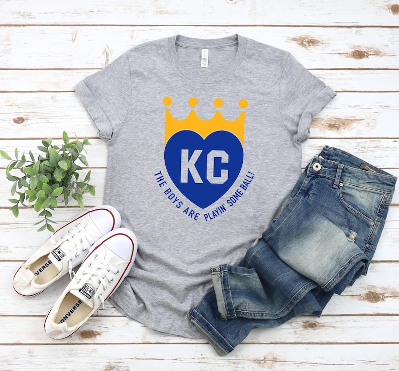 May include: A grey t-shirt with a blue heart and the letters "KC" inside. The heart has a yellow crown on top. The text "The boys are playin' some ball!" is printed below the heart.