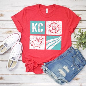 May include: Red t-shirt with a distressed graphic featuring the letters "KC", a soccer ball, stars, and curved lines in white and teal. The shirt is paired with denim shorts and white sneakers on a wooden surface.