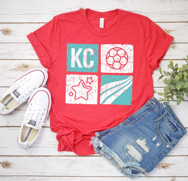 May include: Red t-shirt with a distressed graphic featuring the letters "KC", a soccer ball, stars, and curved lines in white and teal. The shirt is paired with denim shorts and white sneakers on a wooden surface.
