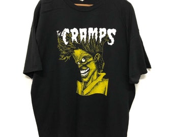 May include: Black t-shirt featuring a yellow and green graphic of a cartoon character with spiky hair and a wide grin. The text "The Cramps" is above the graphic in white. The shirt is hanging on a wooden hanger.