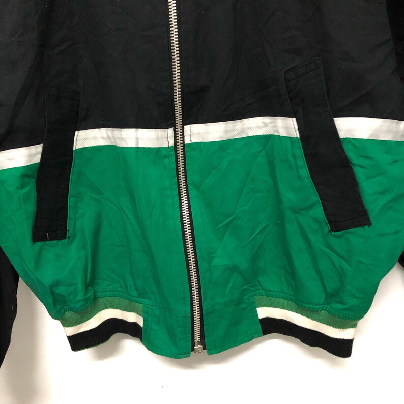 May include: A black, white, and green bomber jacket with a zipper closure. The jacket has a white stripe across the chest and a green bottom with a white and black stripe.