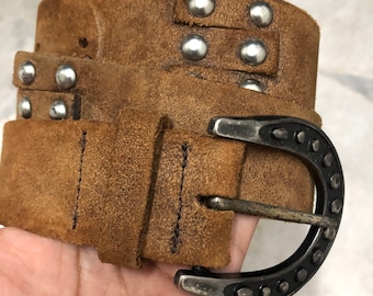 May include: A brown suede belt with a horseshoe-shaped buckle and silver studs. The belt is worn and has a distressed look.