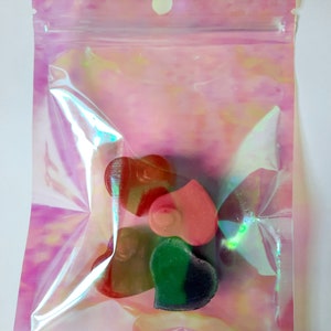 May include: A clear plastic bag with a pink iridescent finish contains four heart-shaped candies. The candies are red, pink, green, and brown.