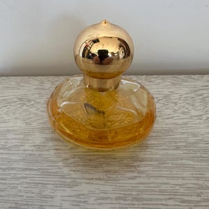 May include: A round, clear glass perfume bottle filled with amber-colored liquid. The bottle features a textured, scalloped design around the base and is topped with a polished, spherical gold-toned cap that reflects the surrounding environment.