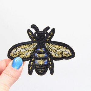 May include: A black and gold embroidered bee patch with blue stripes. The bee has a detailed design with wings and a body.