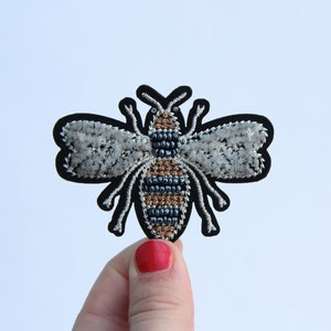 May include: A black and white embroidered bee patch with a brown and black striped body. The bee has a fuzzy texture and is a great addition to any craft project.