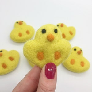 May include: Five yellow felt chicks with orange beaks and black eyes. The chicks are arranged in a semi-circle with one chick in the center being held by a hand.