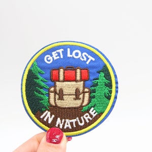 May include: Circular embroidered patch with a blue backdrop and the words "GET LOST IN NATURE". The patch shows a tan backpack with a red top, green trees, and a yellow border. Suitable for outdoor lovers.