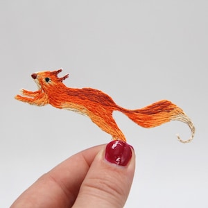 Może przedstawiać: Mała, pomarańczowa wyszywana wiewiórka skacząca w powietrzu. Wiewiórka jest szczegółowo przedstawiona z zakręconym ogonem i małym czarnym okiem.