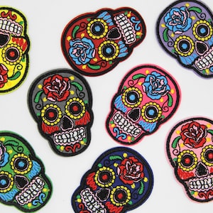 May include: Eight colourful embroidered iron-on patches featuring sugar skulls with floral designs. The skulls are in various colours, including yellow, red, grey, pink, purple, and blue.