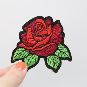 May include: A red embroidered rose with green leaves, outlined in black, is a patch for sewing or ironing onto fabric.